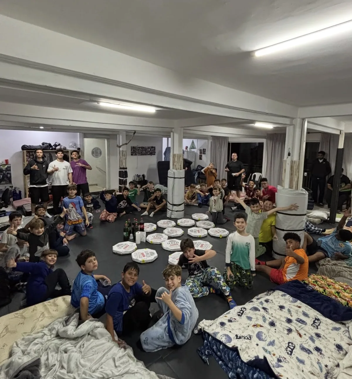 Crianças acampadas no tatame do dojô durante o pernoite, com pizza no centro